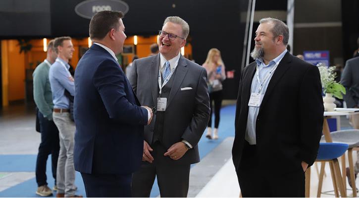three man stand together at a networking event at the Parcel+Post Expo show, with aother attendees in the background and a table with chairs behind them